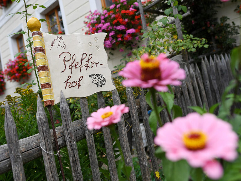 Hinter bunten Blumen und einem Holzzaun zeigt sich das Eingangschild im Garten des Pfefferhofs.