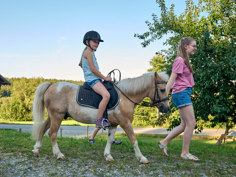Ein Mädchen reitet auf einem Pony, das von einem anderem Mädchen geführt wird beim Pfefferhof.
